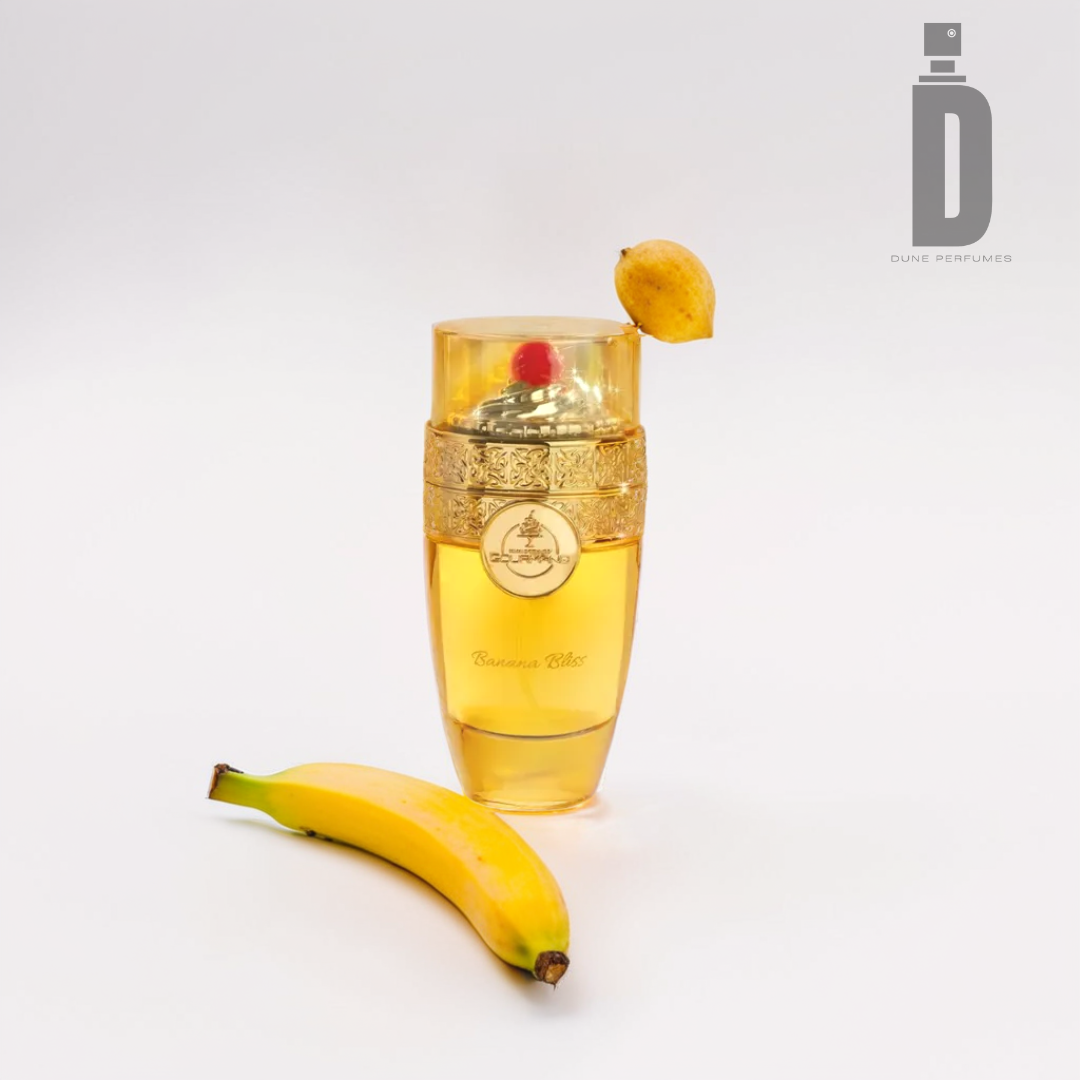 A perfume bottle with an ornate gold design sits beside a ripe banana, topped with a small lemon. Labeled "Banana Bliss 100ml EDP - Paris Corner," it boasts hints of coconut cream. The Paris Corner logo is elegantly displayed against the plain white surface, capturing attention with its exotic allure.