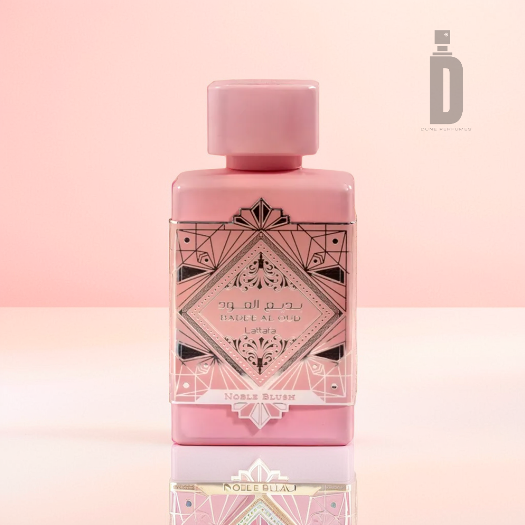 A rectangular-shaped pink perfume bottle, "Badee Al Oud Noble Blush 100ml EDP - Lattafa," featuring an ornate geometric pattern and matching cap, sits gracefully illuminated on a reflective surface. Its presence hints at fragrant notes of rose milk and subtle sandalwood undertones.