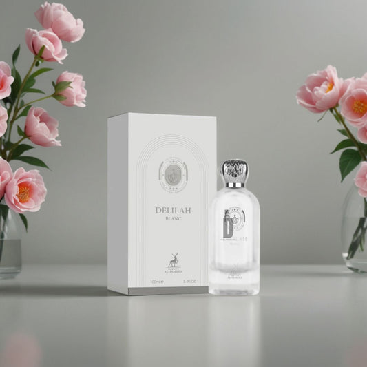 A bottle and box of Maison Alhambra’s Delilah Blanc 100ml EDP sit on a gray surface, with glass vases of pink peonies nearby, emphasizing this luminous fragrance featuring orange blossom notes.