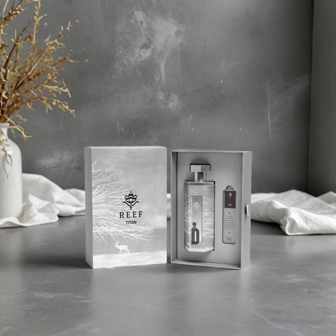 A Titan 200ml EDP by Reef is displayed on a gray surface. The open box reveals the elegant perfume bottle and metallic accessory, with dried plant decor and a white cloth in the background.