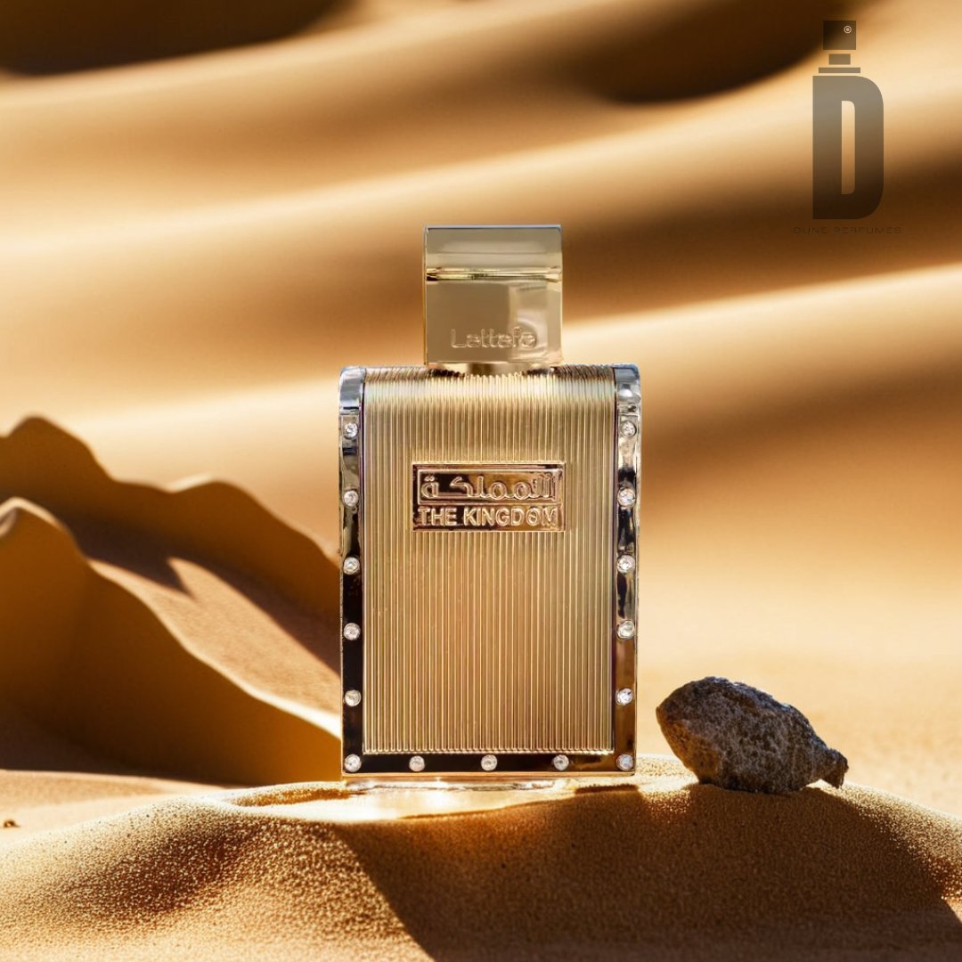 A luxurious golden perfume bottle, labeled "The Kingdom Men 100ml EDP - Lattafa," stands upright on a sandy desert landscape, exuding a masculine fragrance. With its ornate detailing and engraved label from the Lattafa brand, it lends a sensual finish to the scene of small sand dunes and a single rock in the foreground.
