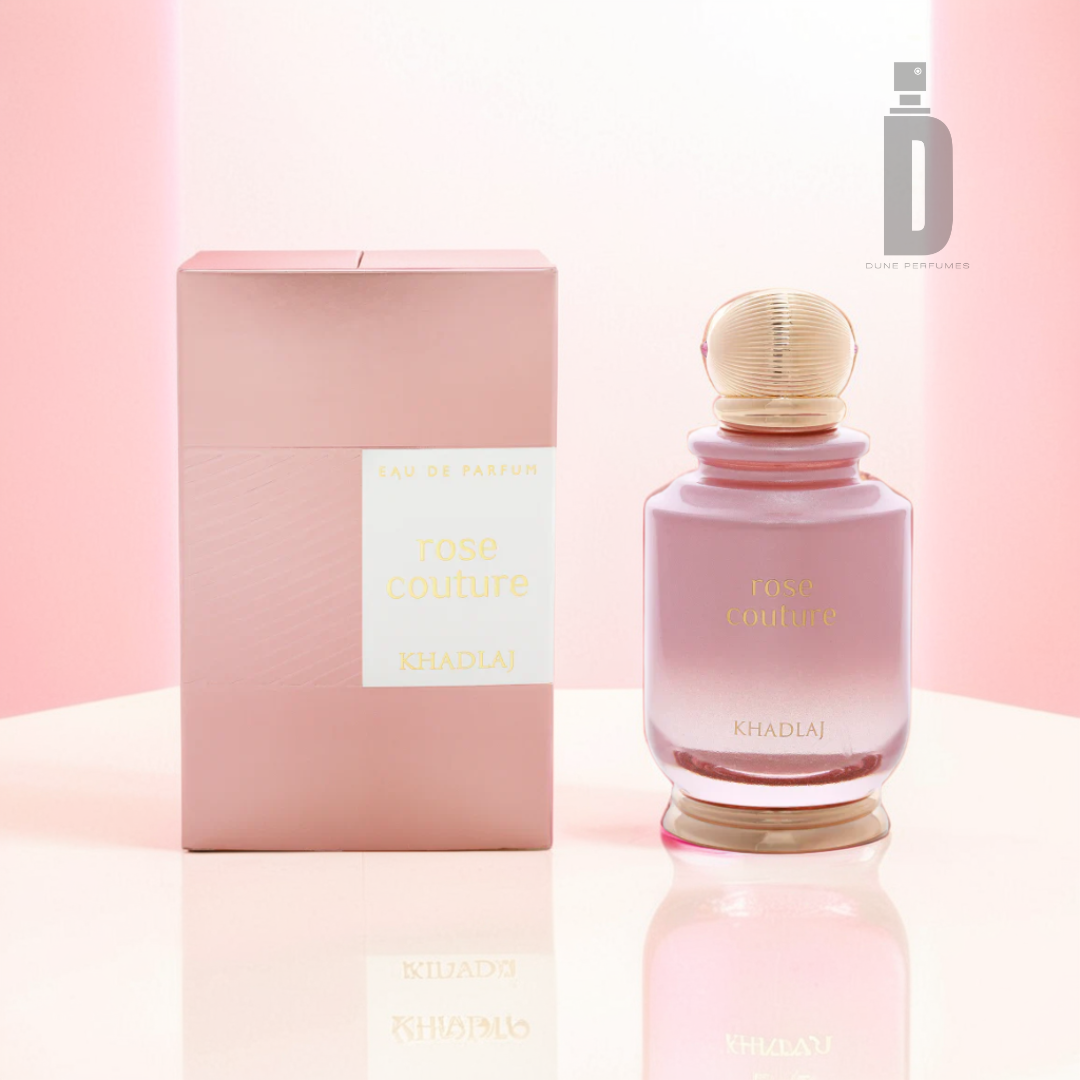 A bottle of Rose Couture 100ml EDP by Khadlaj is placed next to its coordinating pink box. The clear bottle, topped with a golden cap, showcases a subtle Turkish Rose shade. The backdrop features a smooth gradient of light pink and white.