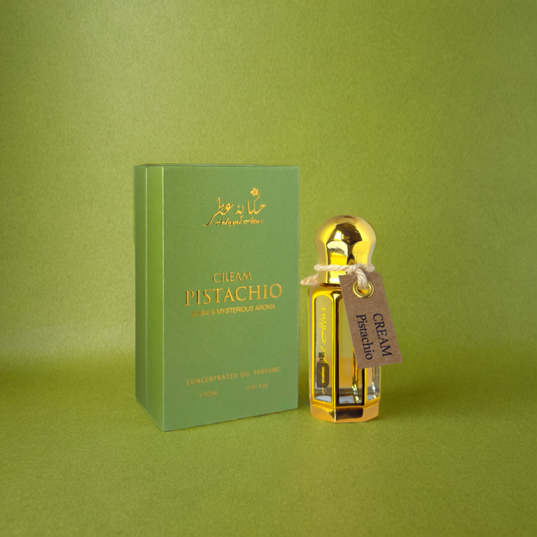A green box labeled "Cream Pistachio" and a small gold bottle, both from Hekayat Attar's Cream Pistachio 12ml perfume oil, are set against a green background.
