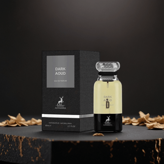 A bottle of Dark Aoud 80ml EDP by Maison Alhambra rests beside its black textured box on a dark surface with dried leaves, evoking rich oud and sandalwood aromas against a deep black background.
