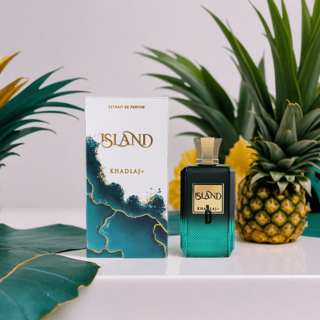 The Island 100ml EDP by Khadlaj stands beside its stylish blue-and-white box, surrounded by tropical leaves and pineapples, evoking a fresh, exotic coconut fragrance.