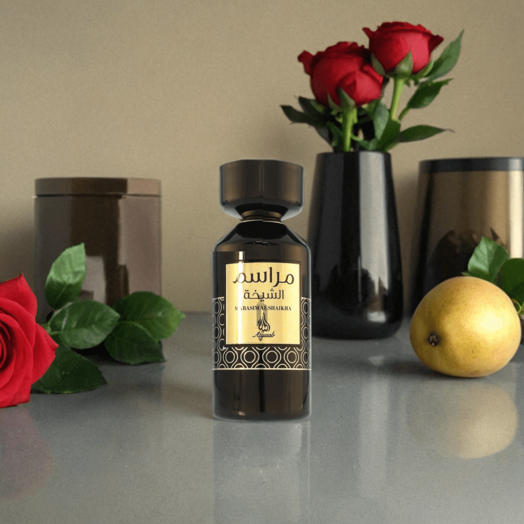 A Marasim Al Shaikha 100ml EDP by Khadlaj stands on a reflective surface, labeled in Arabic, surrounded by a red rose, a pear, two black vases with red roses and green leaves, and a brown jar in the background.