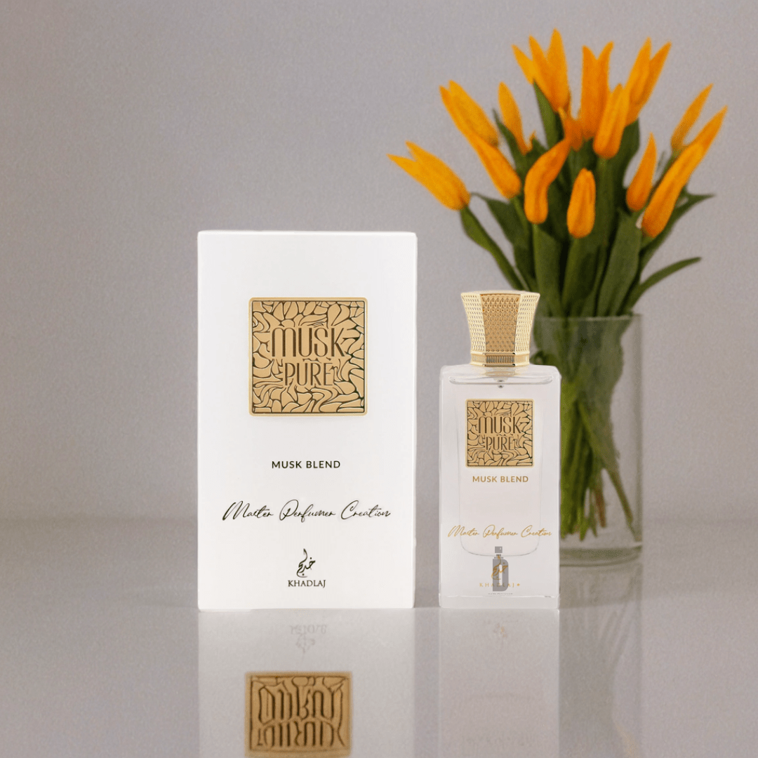 The Khadlaj Musk Pure Musk Blend 60ml EDP bottle and its white and gold box sit on a table, with a clear vase of orange tulips in the background.