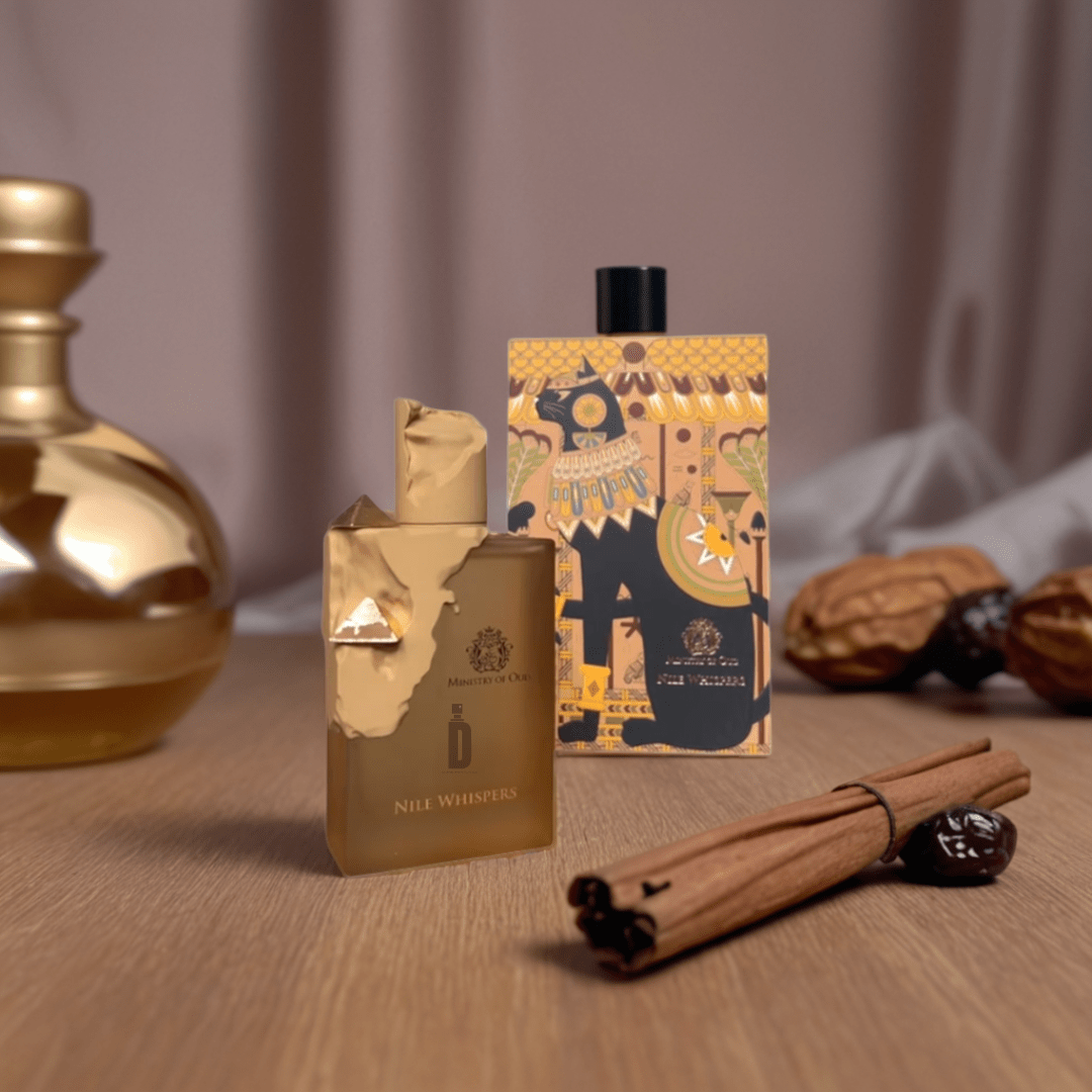 A gold Nile Whispers 100ml EDP by Paris Corner rests beside its vibrant, Egyptian-inspired box. Cinnamon sticks, dates, and a gold vase complete the scene on a soft neutral surface.