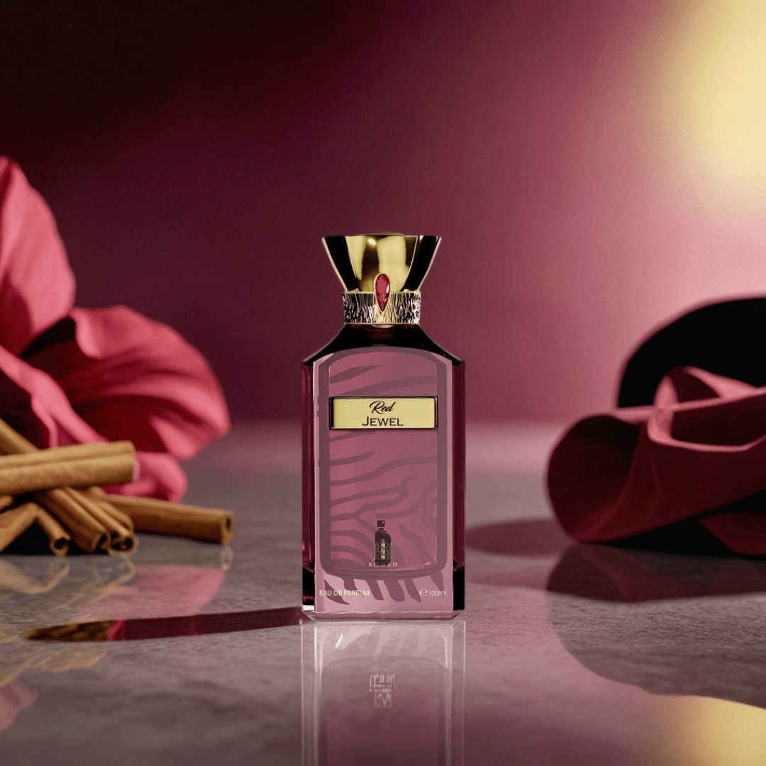 The Red Jewel 100ml EDP by Ahmed Al Maghribi, a luxurious red perfume with a gold cap, rests on marble, surrounded by cinnamon sticks and pink flowers, set against a pink and gold gradient background.