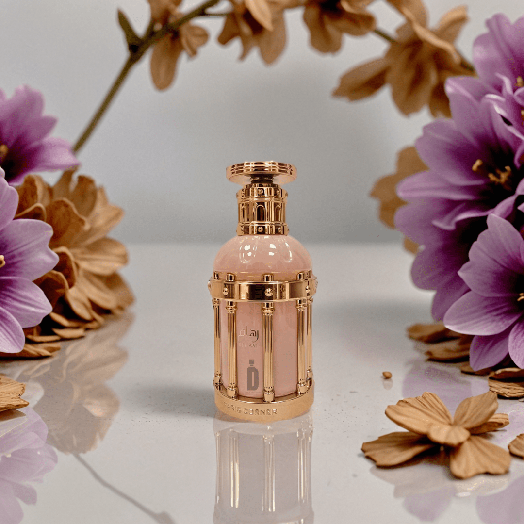 The Reham Rose Petals 100ml EDP by Paris Corner, in a rose gold bottle with a pink center, stands on a glossy surface amid purple and beige flowers against a soft, blurred backdrop.