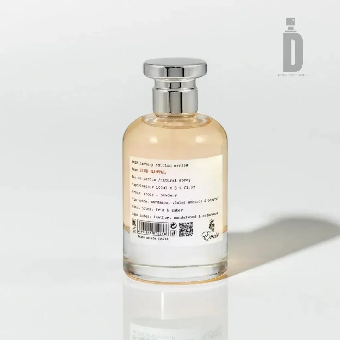 A perfume bottle with a clear glass body and a silver cap stands against a plain white background. The label displays product details in black text, including "Paris Corner Rich Santal 100ml EDP - Emir" and scent notes of "violet, rich santal, leather, and patchouli." This sophisticated fragrance embodies elegance and depth.