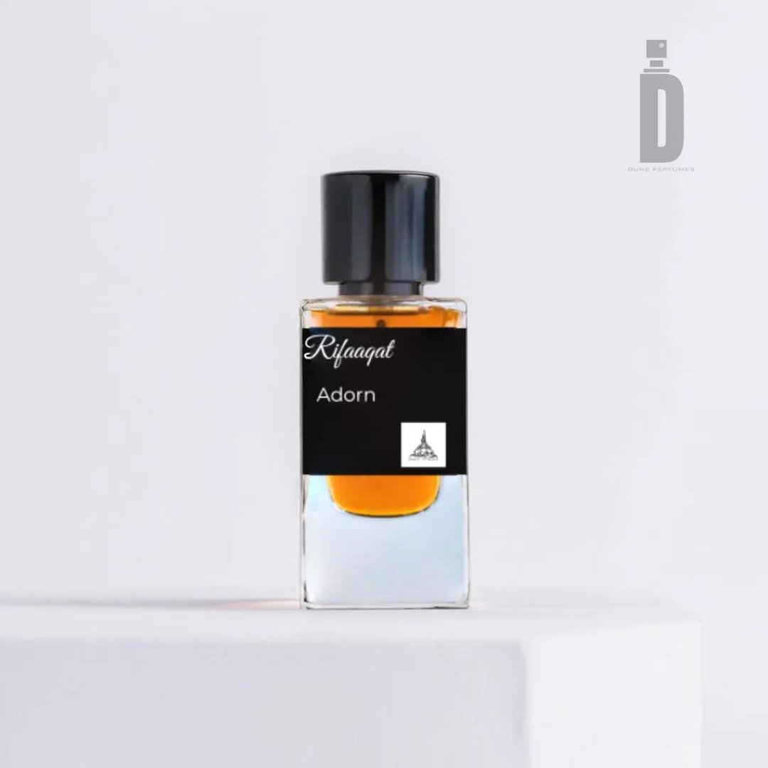 A clear rectangular bottle with a black cap, containing an amber-colored liquid, is displayed against a white background. The label reads "Rifaaqat Adorn 85ml EDP - Paris Corner," highlighting its elegant fragrance and features a small logo at the bottom. The bottle stands on a white platform, exuding timeless beauty.