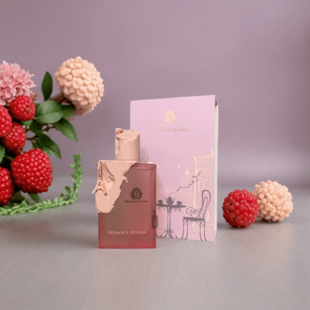 A pink bottle of Romance Reverie 100ml EDP by Paris Corner stands before its matching box, with red and cream flowers arranged to the side on a light gray surface.