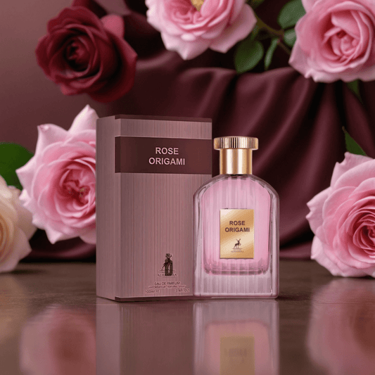 The Rose Origami 100ml EDP by Maison Alhambra is showcased in a pink bottle and box, placed among pink roses and deep red fabric, capturing the luxurious scent of Turkish rose and creamy sandalwood.