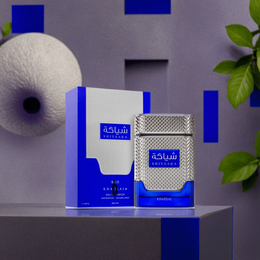 A Shiyaaka Blue 100ml EDP bottle by Khadlaj with silver and blue accents sits on a reflective surface. Its box is nearby, set against gray and blue geometric shapes, green leaves, and a textured white sphere.