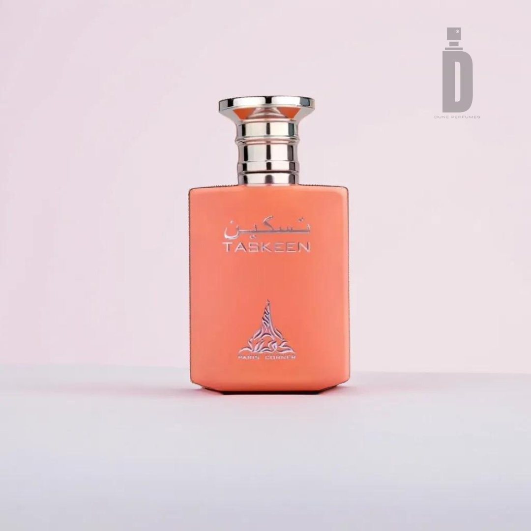 A rectangular perfume bottle labeled "Taskeen 100ml EDP - Paris Corner," featuring a metallic cap and Arabic script, contains a captivating blend within its luxurious fragrance. The soft peach-colored bottle, adorned with an intricate logo at the bottom, is set against a background that transitions gracefully from soft pink to white.
