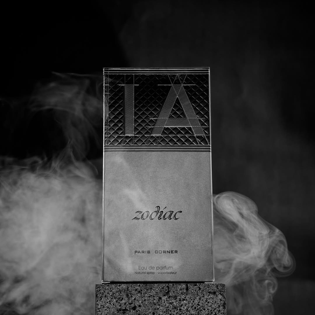 A black and white image features the Zodiac 100ml EDP by Paris Corner atop a concrete block. Smoke swirls around, adding cosmic allure, while the textured background enhances its enticing scent and moody ambiance.