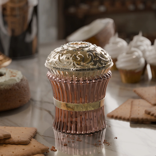 The Creamy Biscuit 100ml EDP by Paris Corner features an ornate, decorative perfume bottle with gold and pink accents on a marble surface, surrounded by biscuits and cupcakes, creating a sweet dessert-themed aesthetic with vanilla fragrance hints.