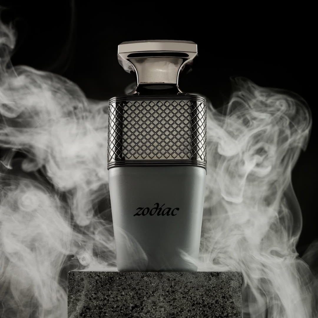 A stylish perfume bottle labeled "Zodiac 100ml EDP - Paris Corner" sits on a stone surface, its sleek metallic grid pattern in the upper part. Mist swirls around, releasing an enchanting scent that creates an elegant, mysterious ambiance with cosmic allure.