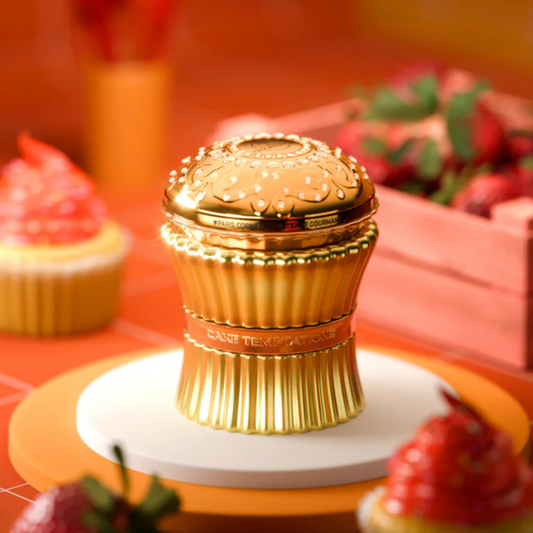 A luxurious, golden Cake Temptation 100ml EDP container by Paris Corner, shaped like a cupcake, sits on a white and orange plate with strawberry-themed decorations. Blurred strawberries in the background invite you to indulge in its delightful gourmand scent.