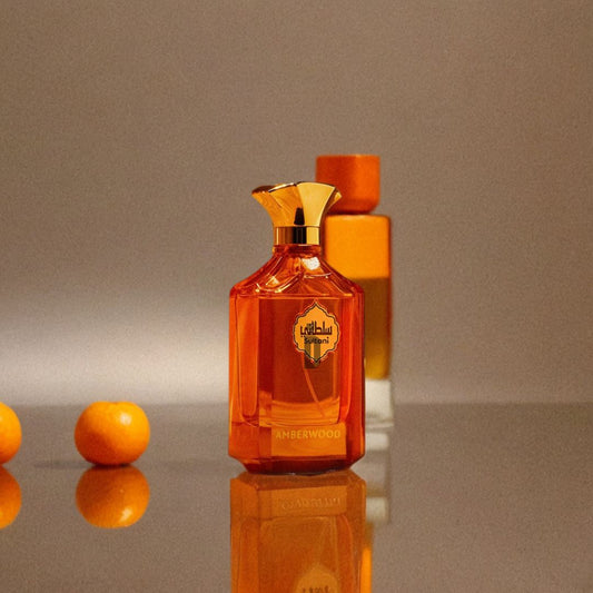 A glossy orange bottle of Sultani Amberwood 100ml EDP by Arabian Oud with a gold cap stands on a reflective surface, exuding luxury. In the softly blurred background are another orange bottle and two small oranges.