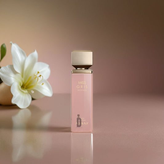 A square, pink bottle of Assaf's Miss Gris Intense 100ml EDP stands on a glossy surface with a white lily, highlighting the bold floral notes and allure of this signature perfume against a softly blurred background.