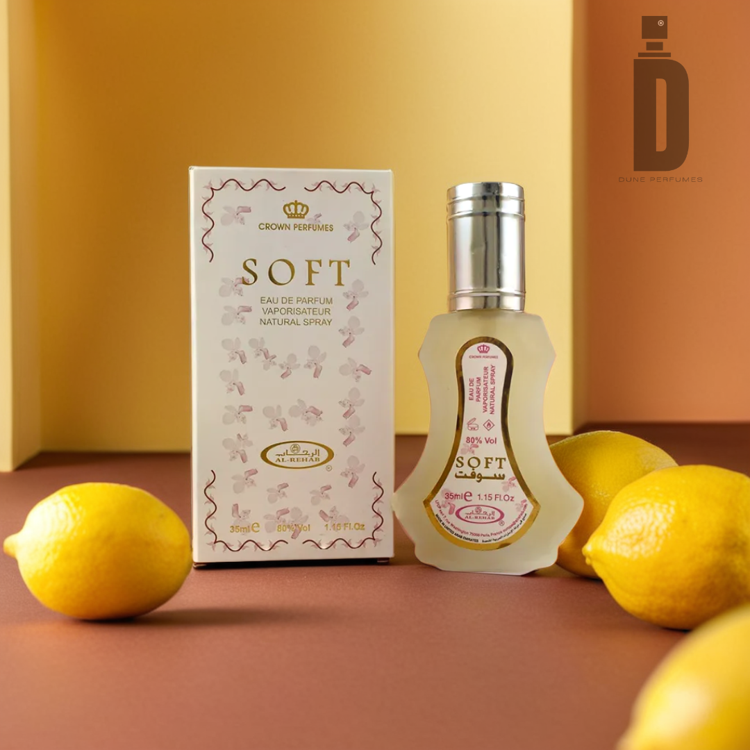 A bottle of Soft - Al Rehab by Al Rehab sits next to its floral-patterned box on a two-toned surface, accompanied by three lemons that impart a fresh, citrusy vibe. Subtle hints of vanilla enhance the fragrance's allure, while the brand logo is visible in the top-right corner.