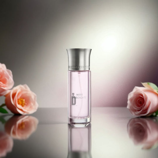 A 200ml bottle of Miss Arrogate EDP by Assaf, with a silver cap, rests on a reflective surface among pink roses and greenery, highlighting its floral fragrance against a softly blurred pink and white background.