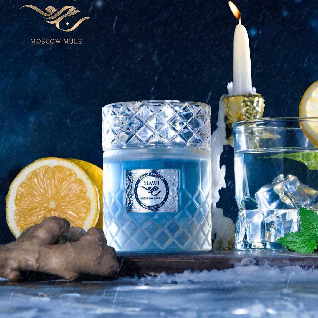 A bottle of "Moscow Mule 100ml EDP" by Paris Corner releases invigorating scents, while surrounded by an arrangement of ice cubes, half a lemon, ginger root, mint leaves, and nestled in a gold candle holder. The dark backdrop embellished with falling snowflakes accentuates the refreshing fragrance of this winter scene.