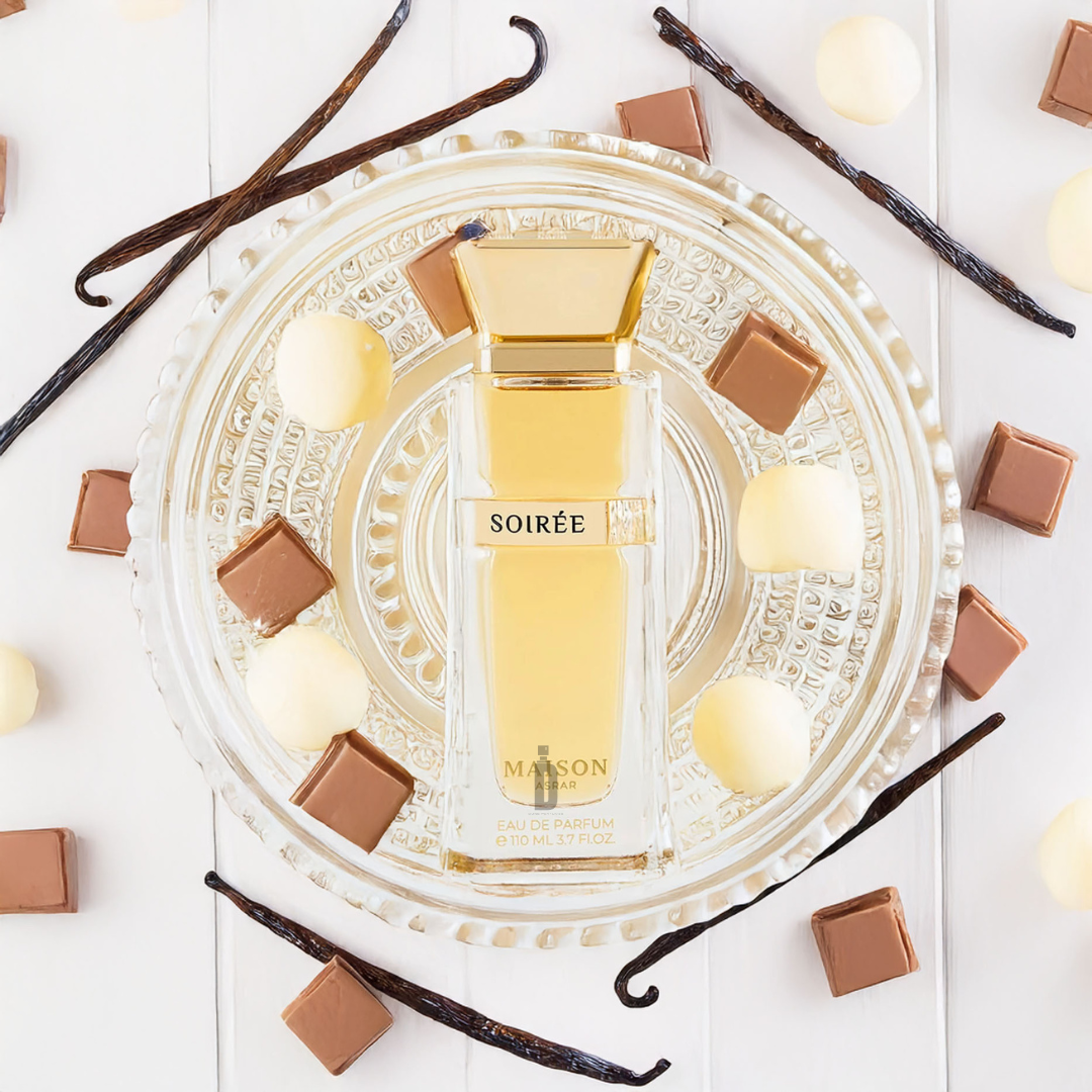 The "Soiree 110ml EDP - Maison Asrar" by Gulf Orchid rests elegantly on an ornate glass plate, embodying a floral bouquet. Surrounded by vanilla pods, white chocolate balls, and milk chocolate squares on a white wooden surface, it creates the perfect evening fragrance.