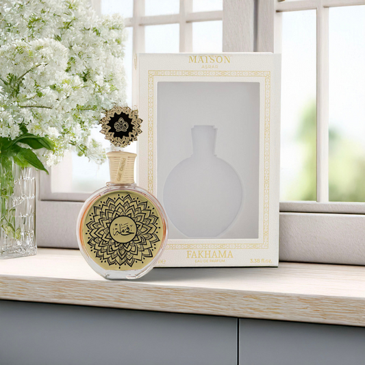 The Fakhama 100ml by Maison Asrar, a Gulf Orchid fragrance, is showcased in an intricately designed bottle with gold patterns. It pairs with its elegant gold-accented box on a wooden surface. A vase of white flowers completes the scene, set against a bright window with a blurred outdoor view.