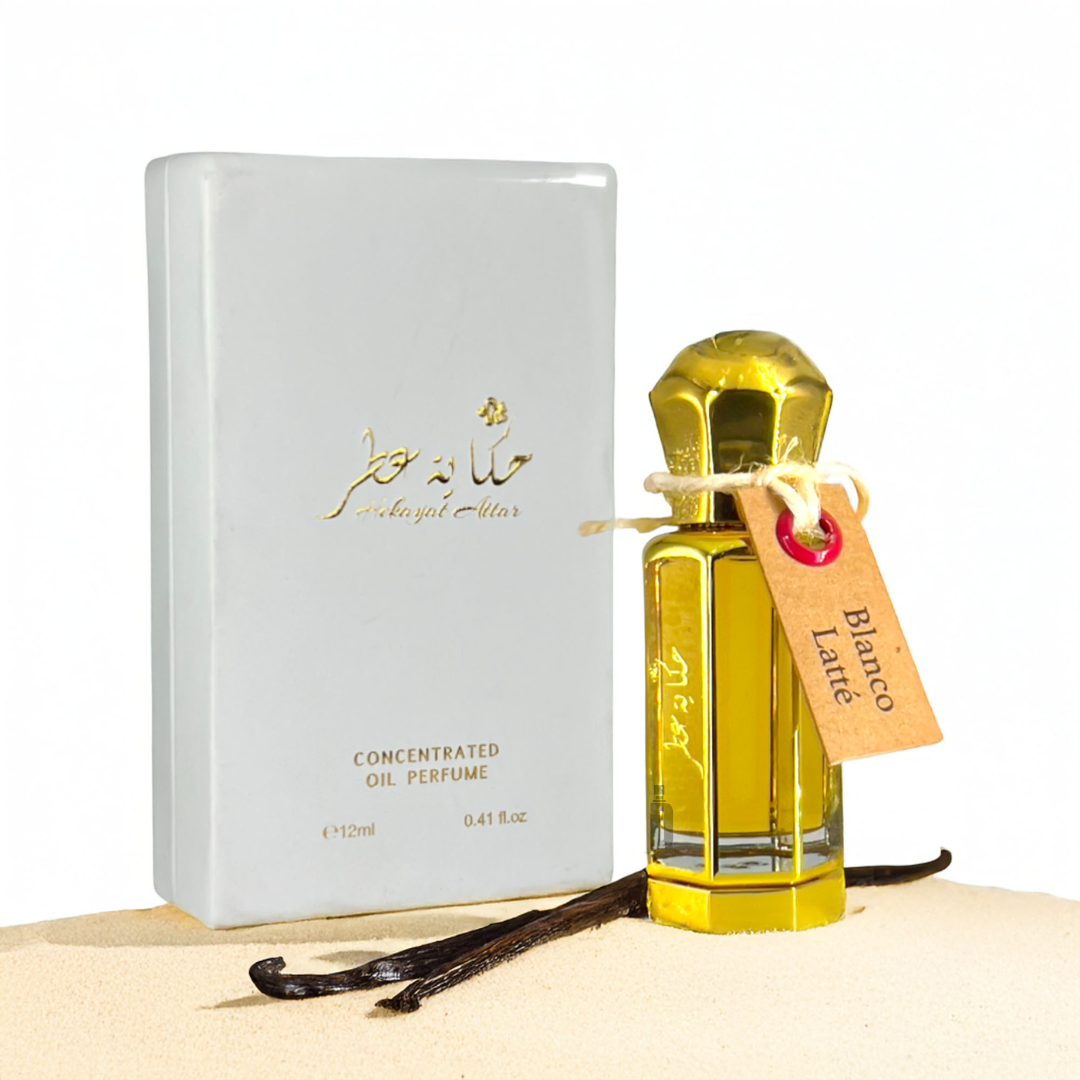 The Blanco Latte 12ml Perfume Oil by Hekayat Attar, in a glass bottle with a gold cap and labeled tag, is displayed next to a white box. Two vanilla pods rest on sand in the foreground, enhancing the Creamy Caramel Vanilla scent.