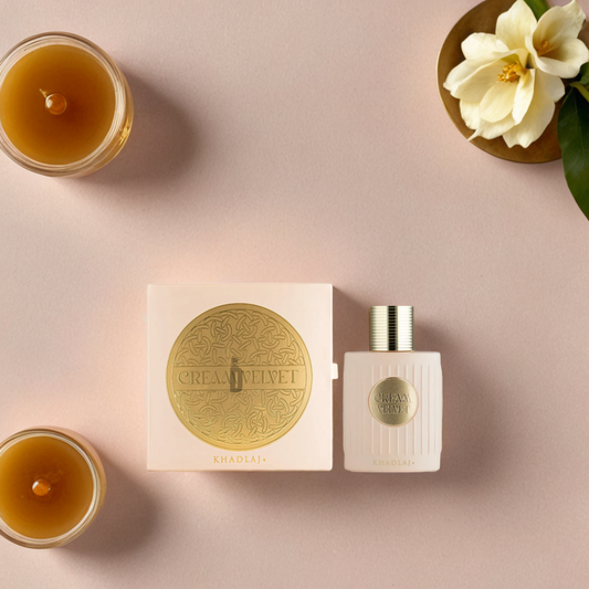 The elegant Cream Velvet 100ml EDP by Khadlaj is set against a soft pink background. Its luxurious bottle and box are accompanied by two lit candles and a white flower with green leaves, creating a graceful scene with a velvety finish.