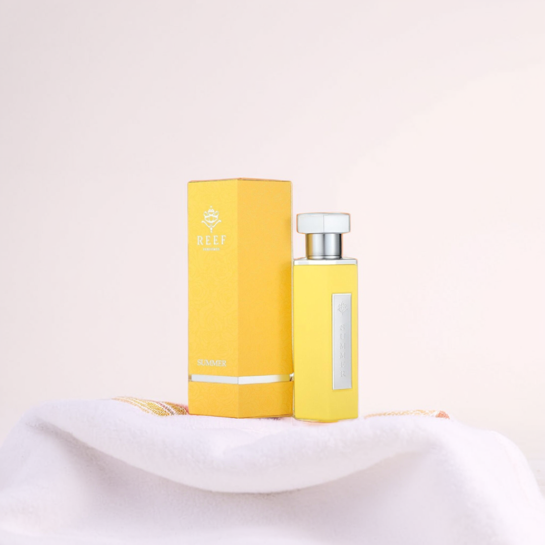 The Summer Yellow 100ml EDP by Reef, featuring hints of orange blossom, comes in a yellow bottle with a matching box. It sits on a white textured surface against a soft pink background.