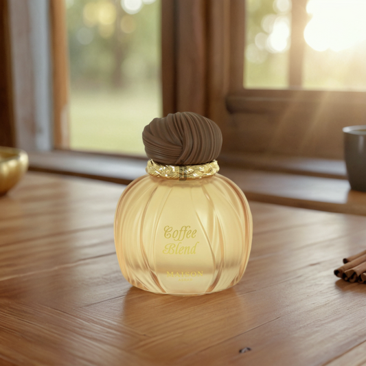 A beautifully designed bottle of Maison Asrar's "Coffee Blend" 100ml EDP by Gulf Orchid sits on a wooden table near a window. Adorned with a brown cap and golden band, it boasts rich fragrance notes as warm sunlight delicately illuminates the scene.