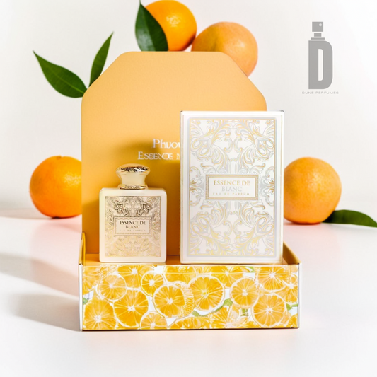 The Essence de Blanc 100ml EDP from Fragrance World features a perfume set with a patterned bottle and matching box, both adorned with citrus designs that enhance the fragrance's allure. Fresh oranges and green leaves surrounding the set add a vibrant touch, while a hint of ambergris elevates its complexity. A minimal logo is visible in the background.