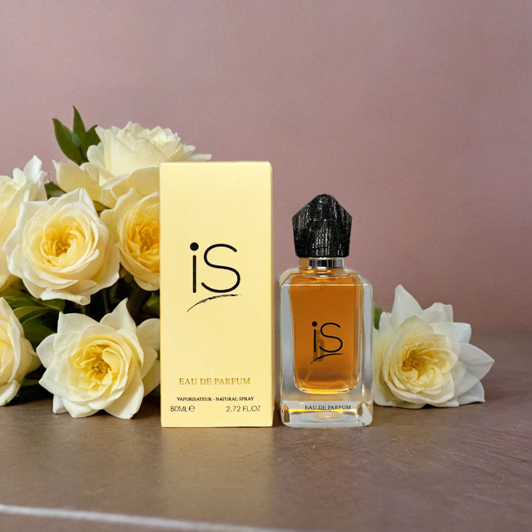 The Is 75ml EDP by Fragrance World sits alongside its cream-colored box, surrounded by cream and yellow roses—evoking Rose de Mai notes—on a light brown surface with a soft mauve background.