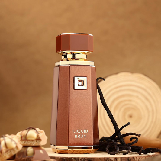 A sophisticated brown perfume bottle of "Liquid Brun 100ml EDP - Fragrance World" stands upright, adorned with gold accents. Surrounded by bourbon vanilla pods and small confections, it exudes warmth against a warm wooden backdrop.