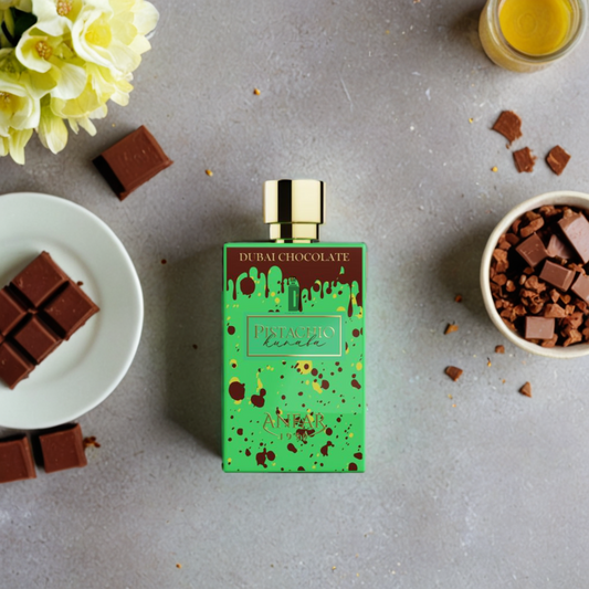 A green bottle of Anfar's Dubai Chocolate Pistachio Kunafa 80ml EDP rests on gray, surrounded by chocolate, crushed chocolate in a bowl, a glass of pale yellow liquid, and yellow flowers—capturing the essence of Middle Eastern desserts.
