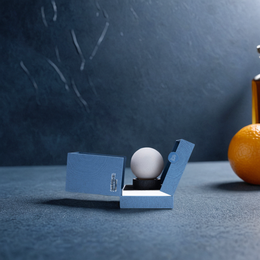 Cafe Citadel 100ml EDP by Aurora Scents is displayed in a blue ring box on a textured blue surface, exuding smoky elegance, with an orange and a glass bottle softly blurred in the background.