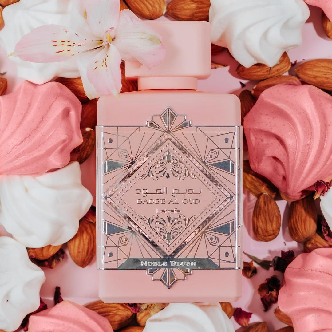 The "Badee Al Oud Noble Blush 100ml EDP" by Lattafa, in its ornate geometric pink bottle, is gracefully encircled by almonds, pink meringues, rose petals, and a white lily on a pink background. Notes of sandalwood evoke an essence akin to sweet rose milk and the Noble Blush experience.