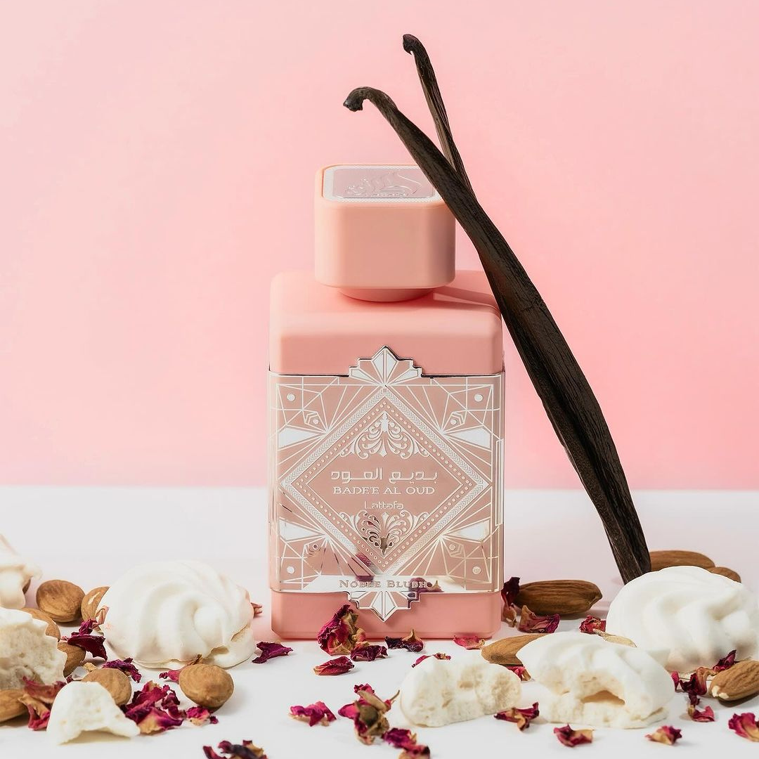 A pink perfume bottle labeled "Badee Al Oud Noble Blush 100ml EDP - Lattafa" rests on a light pink background, surrounded by vanilla pods, nuts, meringues, and scattered rose petals. This setting creates an elegant and aromatic presentation.