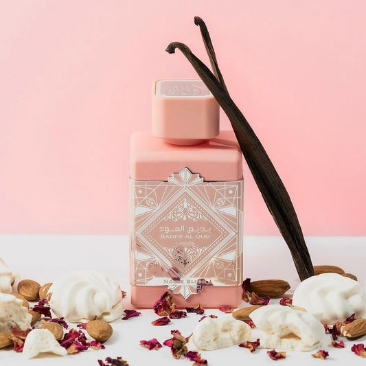 A pink perfume bottle labeled "Badee Al Oud Noble Blush 100ml EDP - Lattafa" rests on a light pink background, surrounded by vanilla pods, nuts, meringues, and scattered rose petals. This setting creates an elegant and aromatic presentation.
