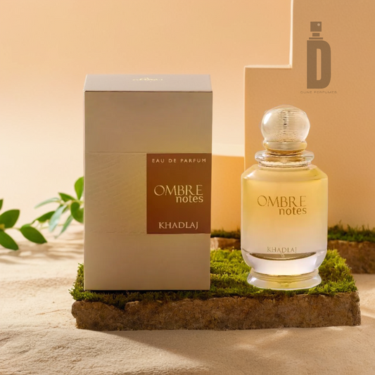 The "Ombre Notes 100ml EDP" by Khadlaj, contained in a sleek glass bottle, is showcased alongside its stylish box. These items are arranged on a textured surface adorned with moss and leaves, evoking an earthy and natural atmosphere.