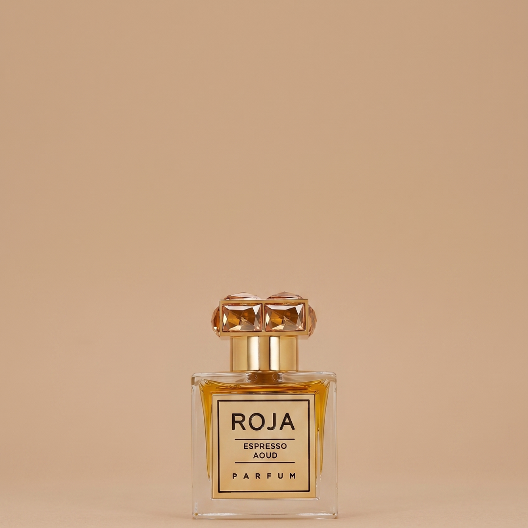 A 100ml bottle of Roja Parfums Espresso Aoud EDP stands centered on a beige background; the clear glass reveals amber liquid, suggesting its rich oud and gourmand scent, finished with a gold, gem-set cap.