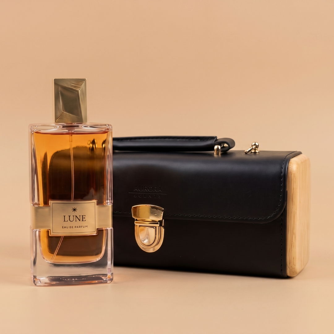A 100ml bottle of Aurora Scents Lune Eau de Parfum, featuring a gold cap and label, sits beside a black leather clutch with a gold clasp on a beige background.