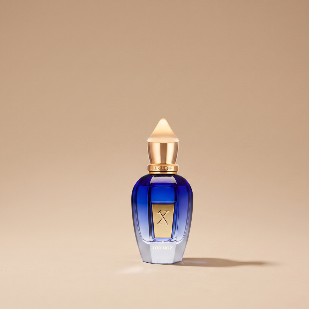 A blue glass bottle of Xerjoff Torino 21 100ml EDP, featuring a gold cap and label, stands upright on a beige background with a soft shadow to the right, evoking the fresh elegance and energetic spirit of this aromatic citrus fragrance.