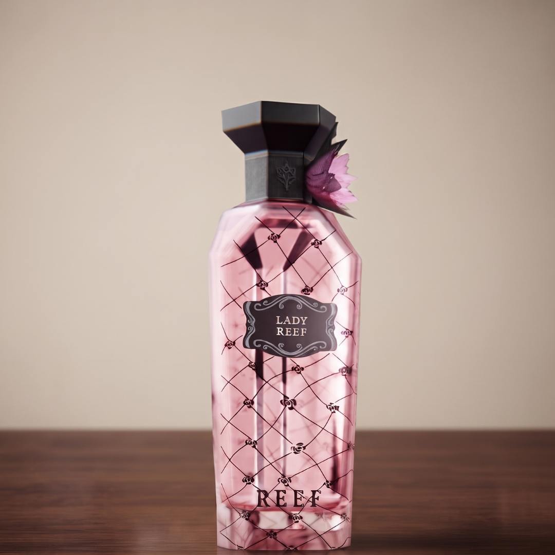 A pink, faceted perfume bottle with a black cap and flower decoration—labeled "Lady Reef 150ml EDP" by Reef—rests on a wooden surface against a plain beige background, beautifully reflecting the refined, feminine essence of this elegant floral fragrance from Reef.