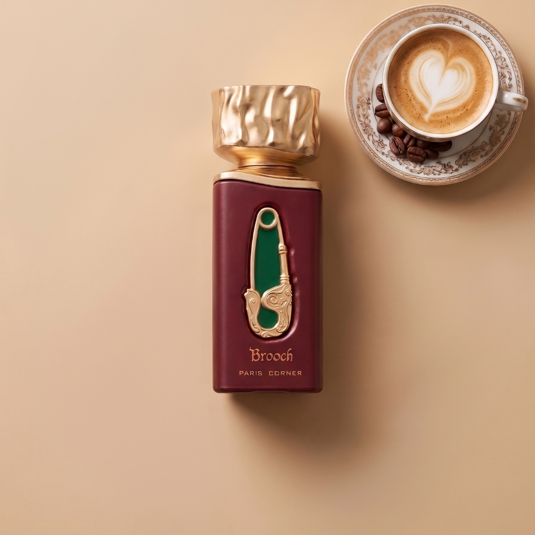 A bottle of Brooch 100ml EDP by Paris Corner, featuring a gold top and green label, rests beside a cup of cappuccino adorned with heart-shaped foam art on a beige surface, capturing the essence of creamy coffee notes and sweet spices in this delectable gourmand fragrance.