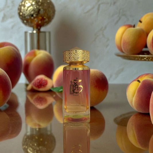 The Khair Peach Delulu 100ml EDP by Paris Corner stands on a reflective surface with fresh peaches and figs, radiating a rich fruity aroma. A gold cap, decorative piece, and fruit plate add elegance to the scene.
